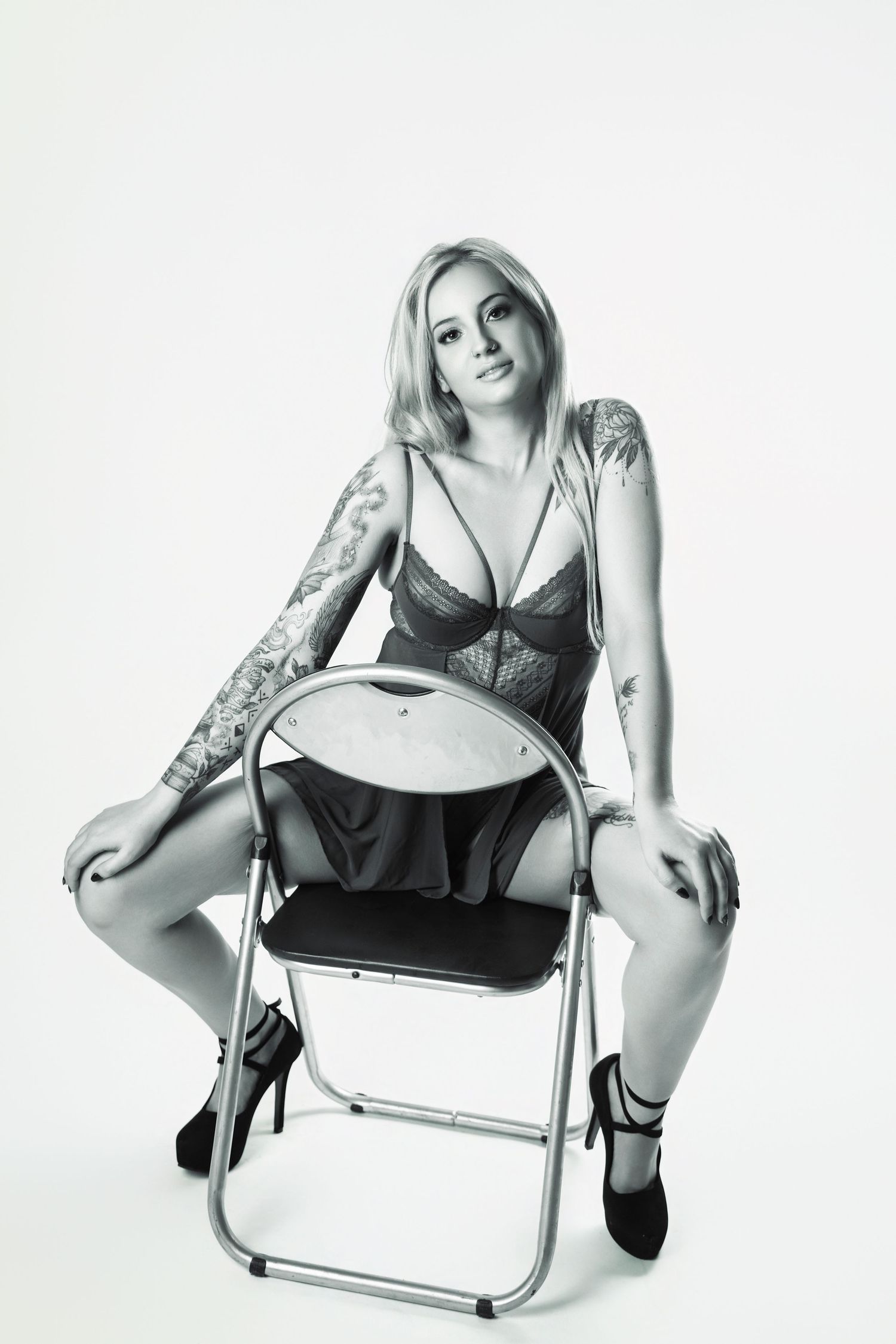 Tattooed blonde woman in black lingerie sitting on folding chair in black and white studio photo.