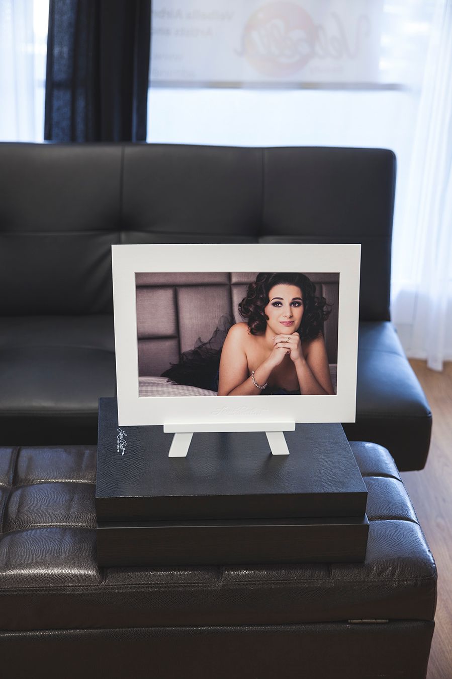 Framed boudoir portrait photo displayed on easel atop black box near leather sofa.