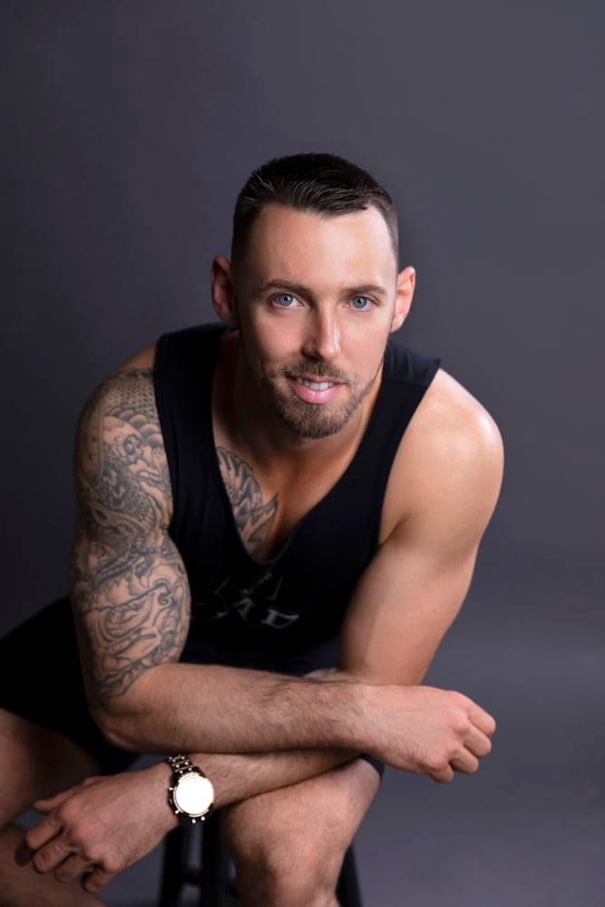 Tattooed muscular man in black sleeveless shirt posing in studio with grey background.