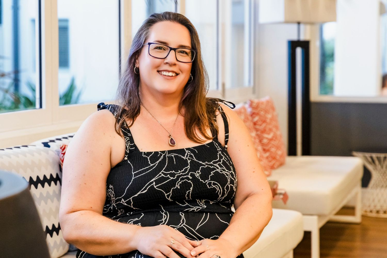 Cheerful woman with glasses in floral top sits relaxed smiling in bright modern interior space.