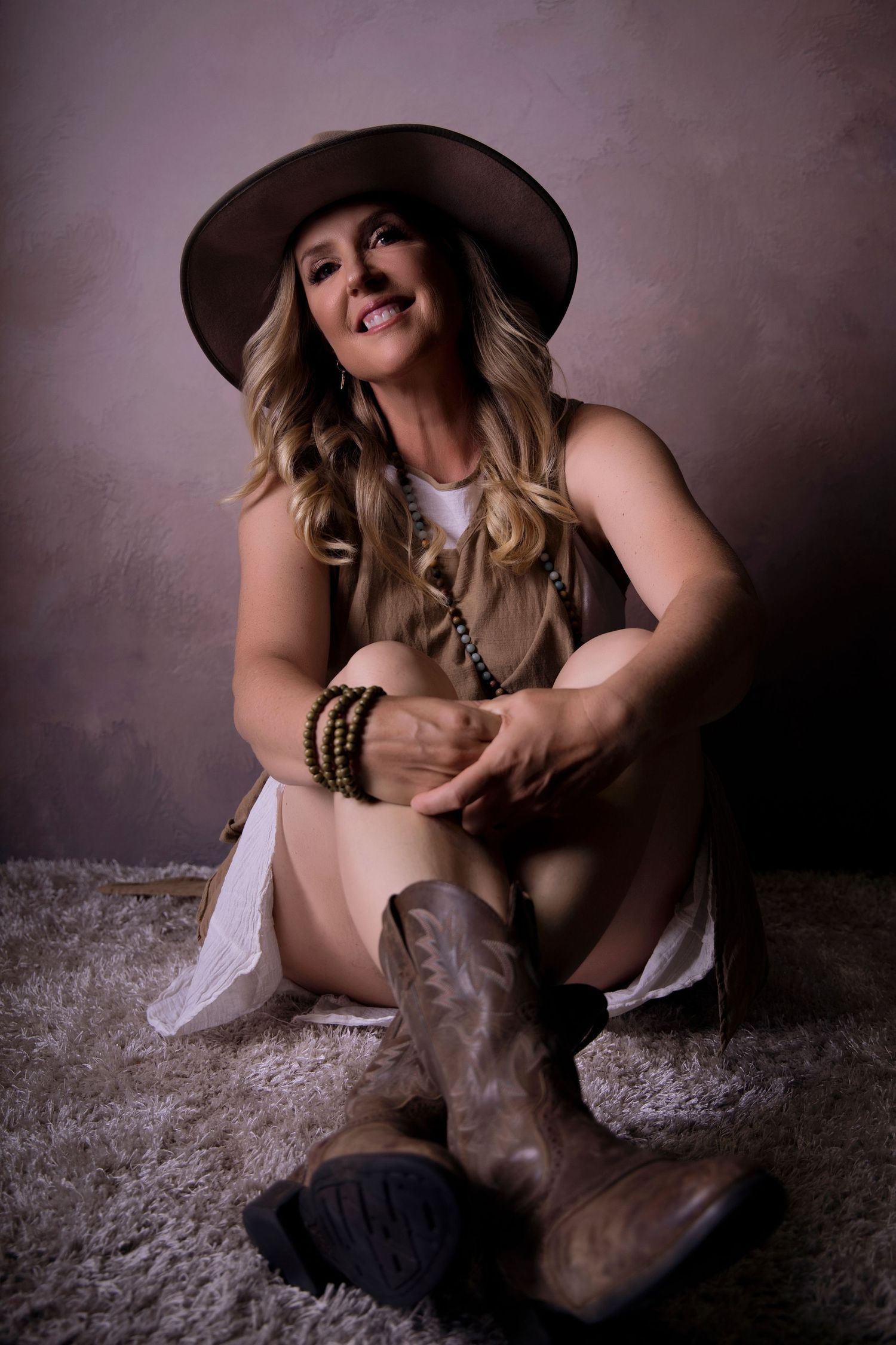 Smiling blonde woman in cowboy hat and boots sitting on fur rug in studio.