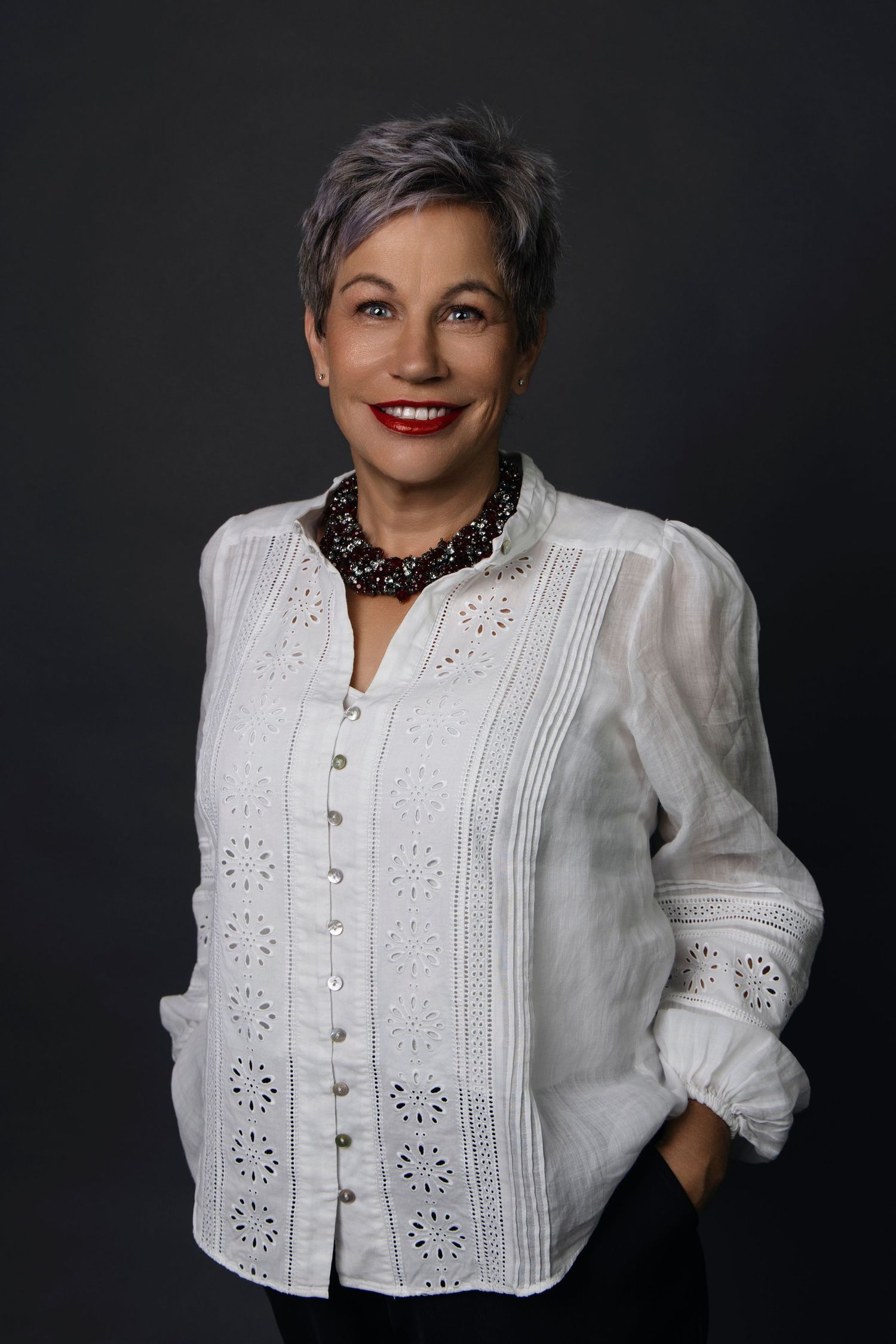 Elegant mature woman with short gray hair wearing white linen blouse and beaded necklace against dark background.