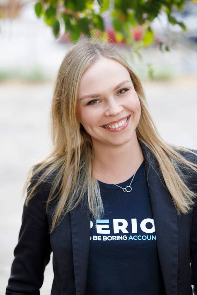 Smiling blonde woman in Perio branded t-shirt and blazer posing outdoors under green trees.