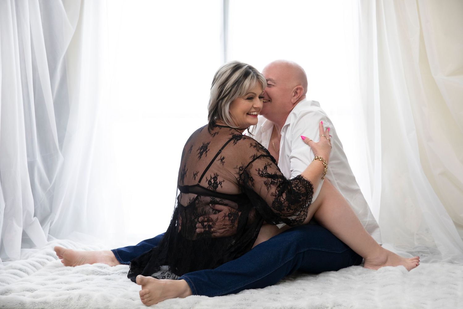 A couple sits intimately on a white fluffy rug, the man kissing the woman's cheek as she smiles warmly.