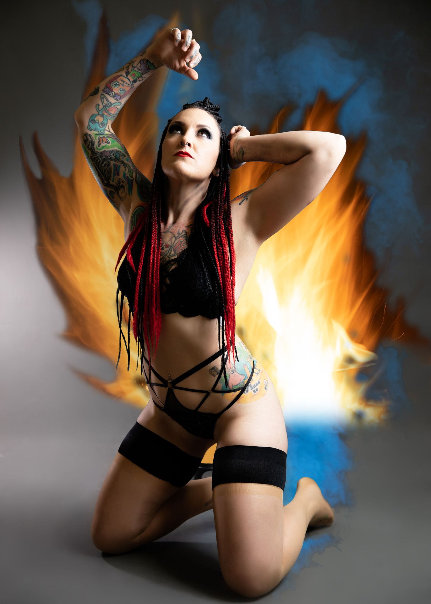 Tattooed woman with red dreadlocks kneels in lingerie before dramatic blue and orange flames.