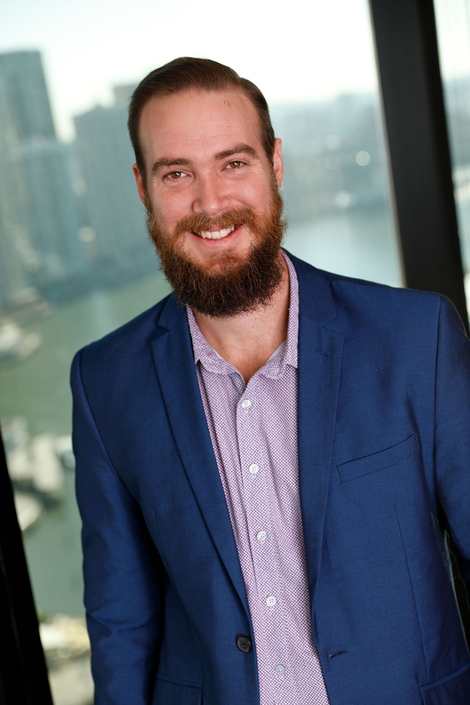 Smiling bearded man in blue blazer with city waterfront background in professional portrait.