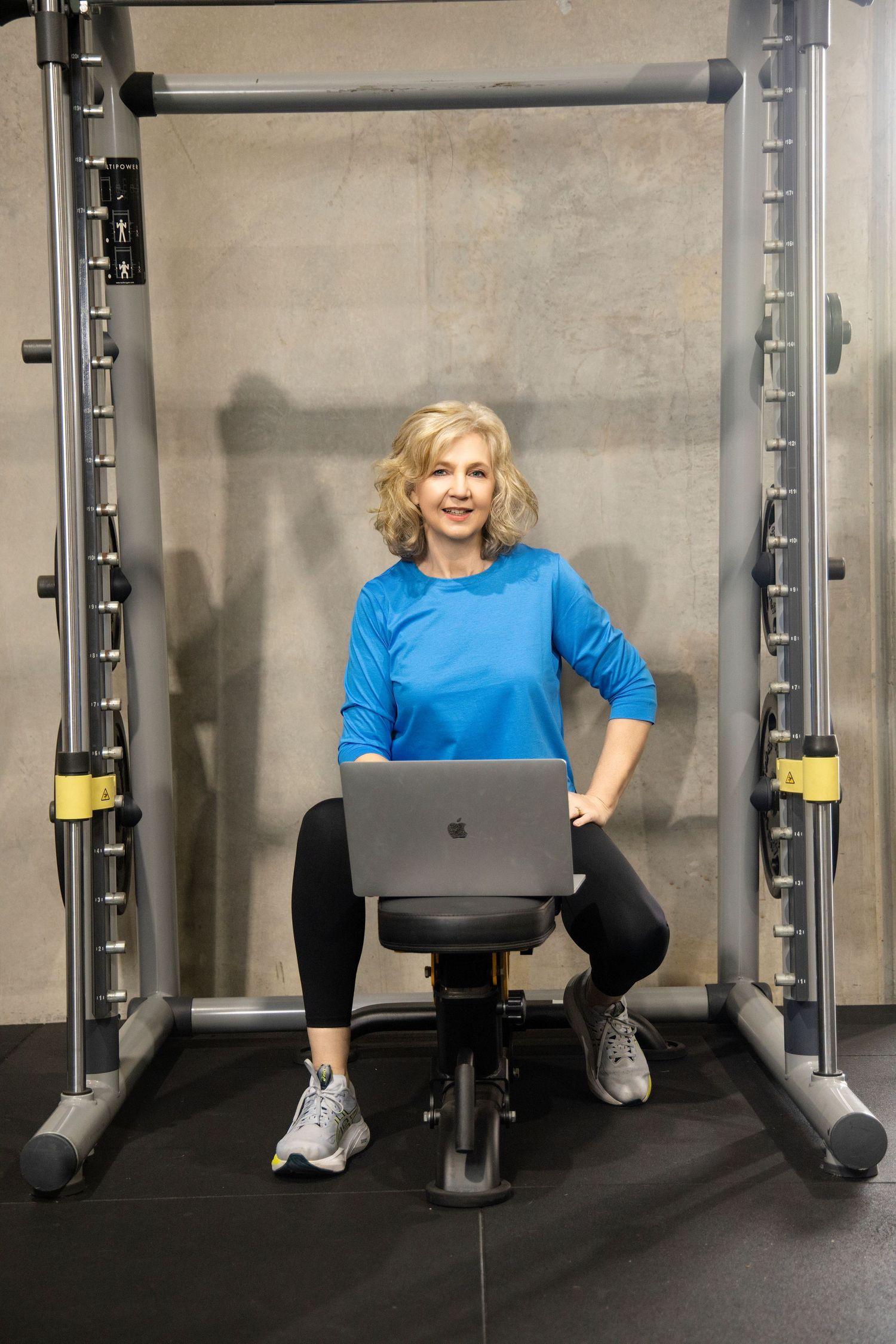 Woman in blue top sits on gym bench with laptop in front of cable machine smiling at camera.