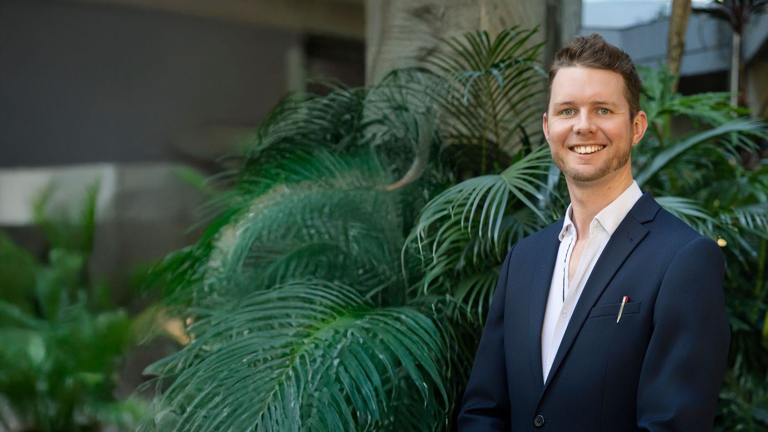 Real estate agent in navy blazer standing outdoors surrounded by lush tropical green plants.