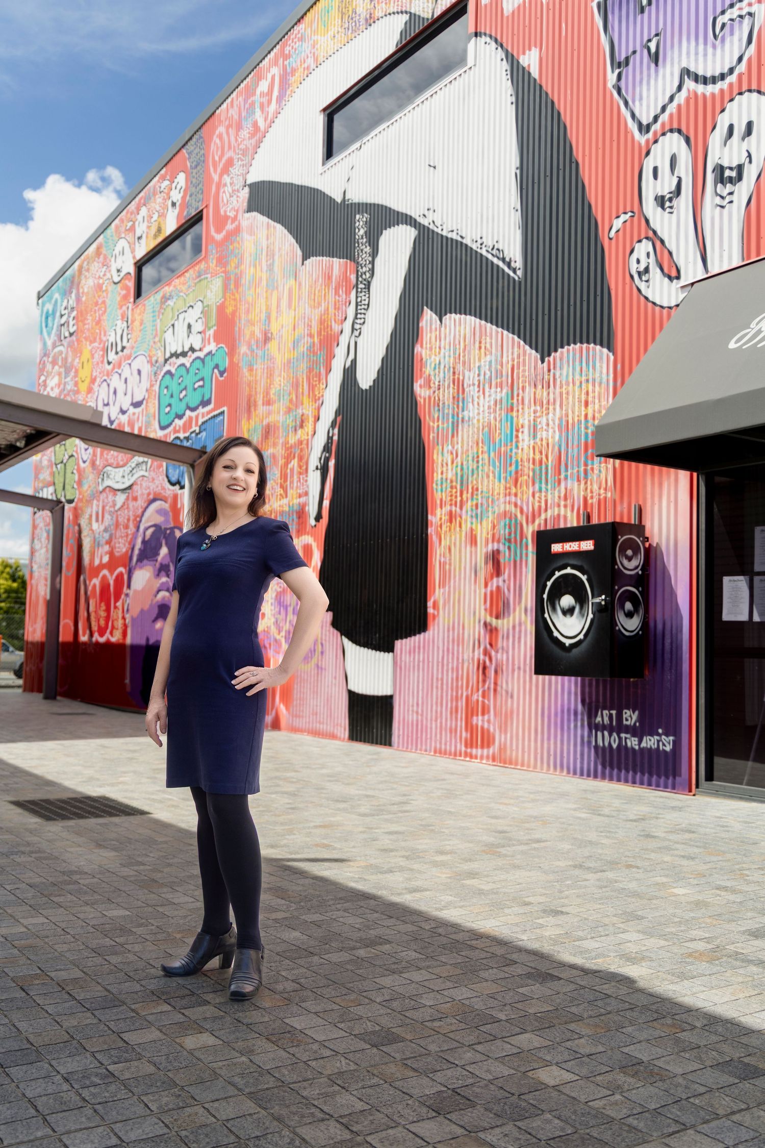Smiling woman in navy blue dress posing in front of colorful urban street art mural on bright day.