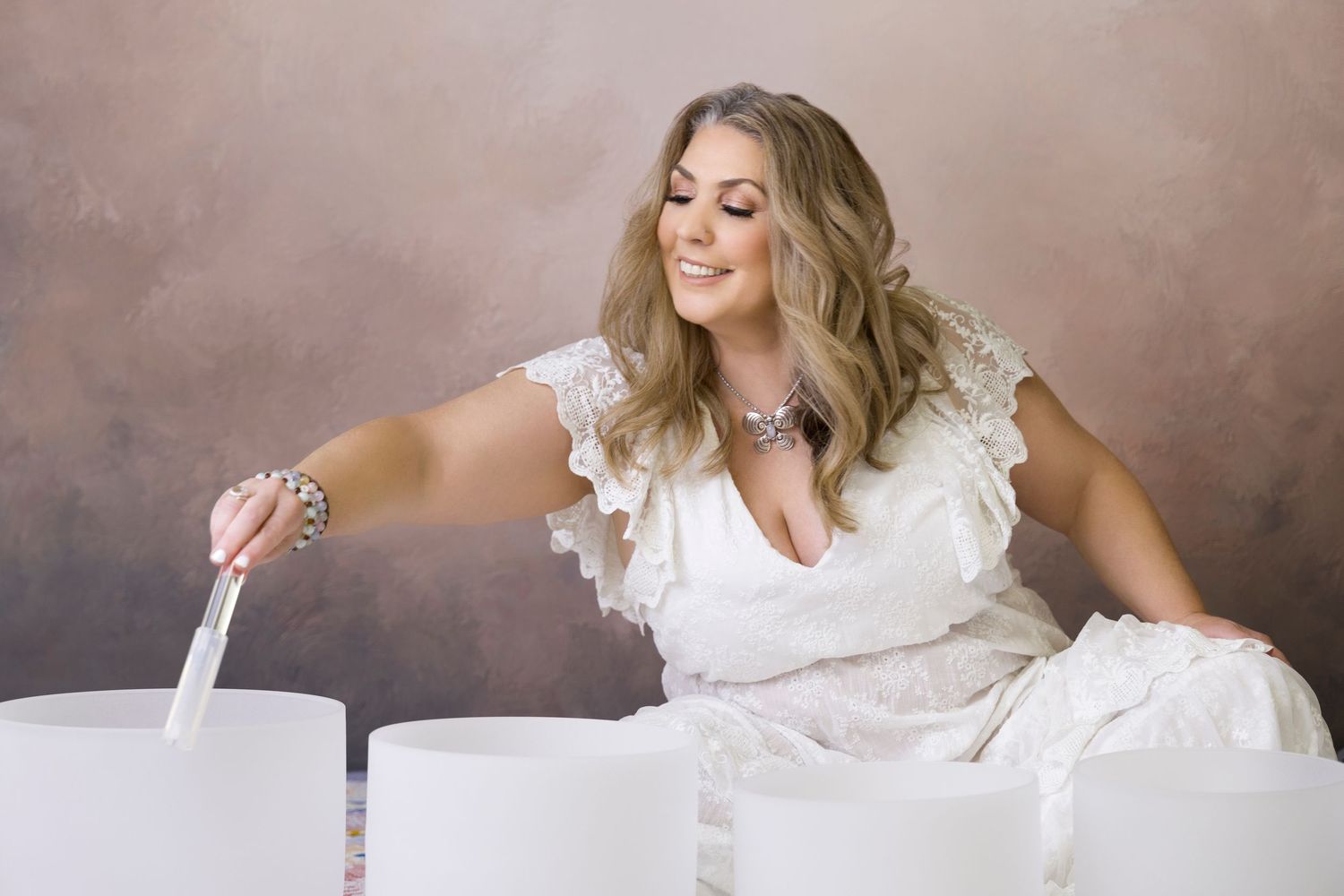 Woman in white lace dress playing crystal singing bowls in a professional studio setting.