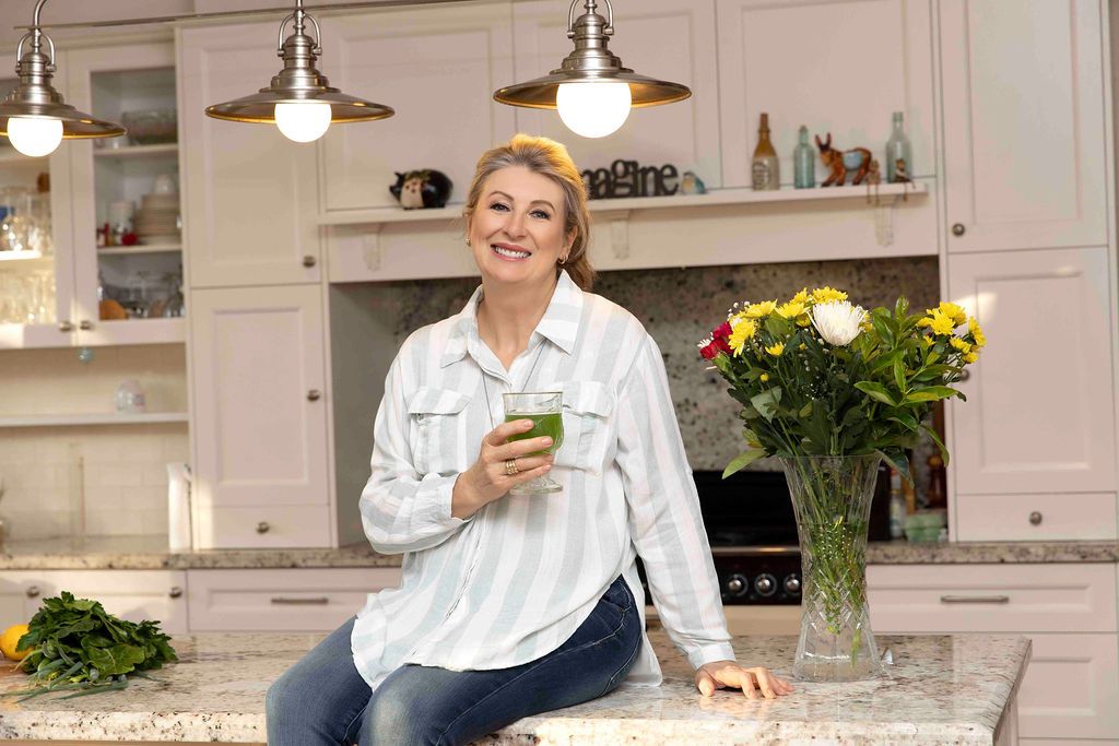 Smiling woman holding green smoothie sitting on kitchen counter with fresh flowers.