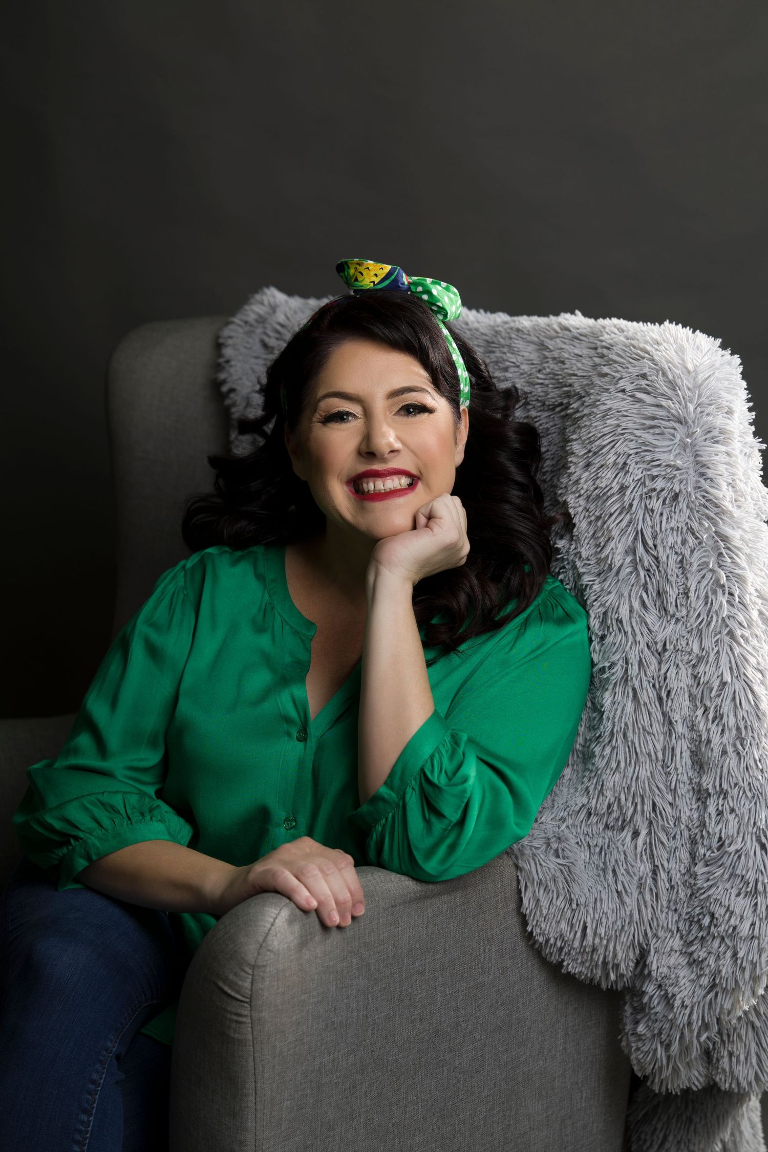 A smiling dark-haired woman in a green blouse sits relaxed in a gray armchair.
