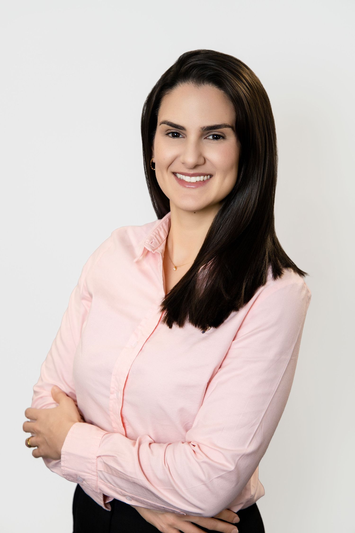 Young brunette businesswoman in pink shirt smiles with arms crossed against white background.