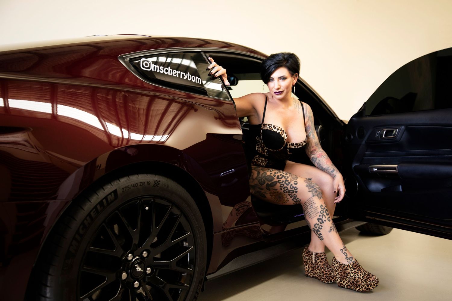 Tattooed woman in black lingerie leaning against dark red car door in glamour portrait.
