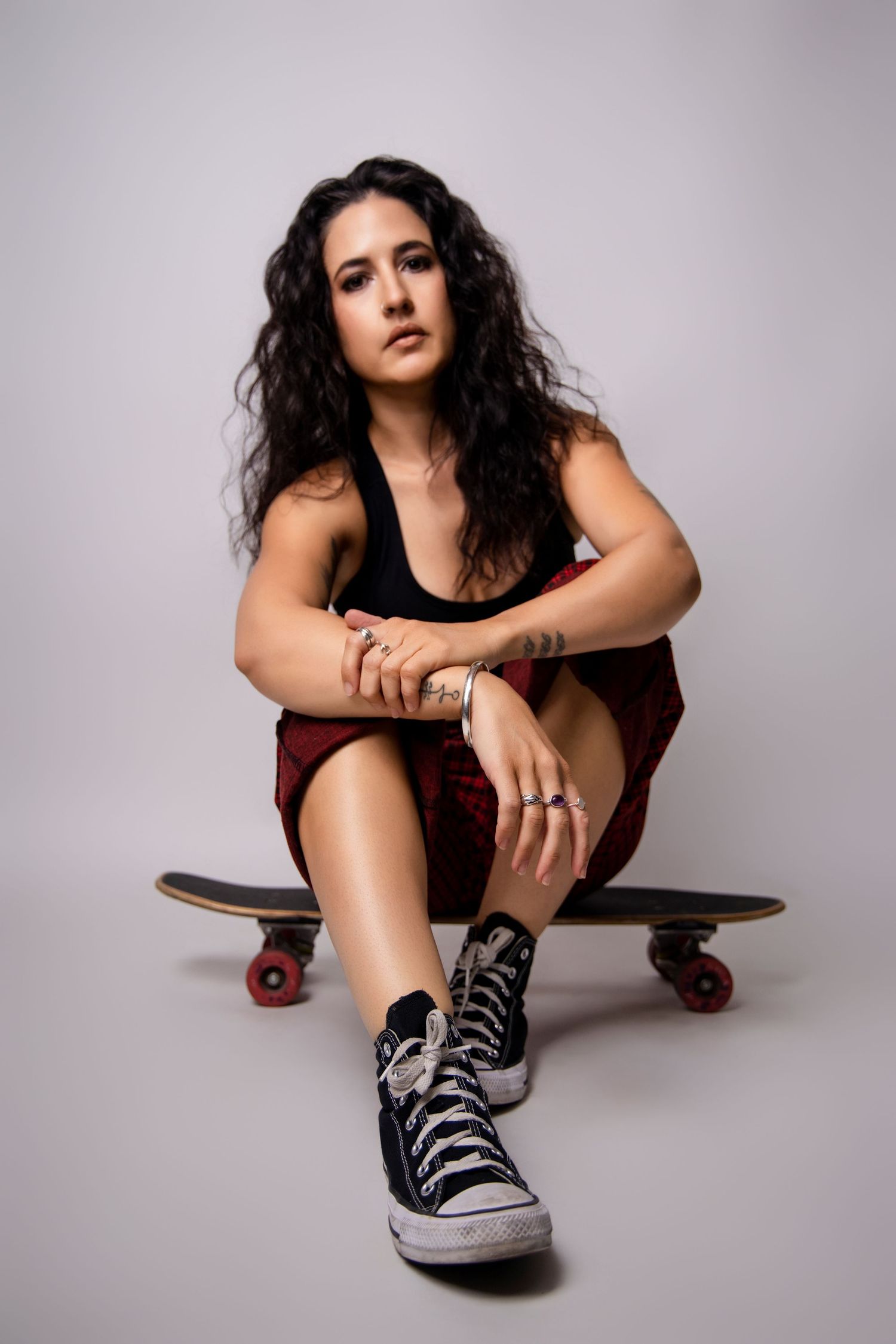 Tattooed young woman sitting on a skateboard wearing Converse sneakers and red plaid skirt in studio.