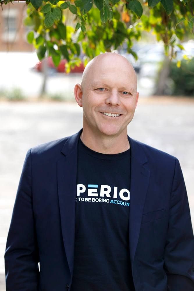 Confident bald man in navy blazer and Perio t-shirt smiling outdoors under green foliage.