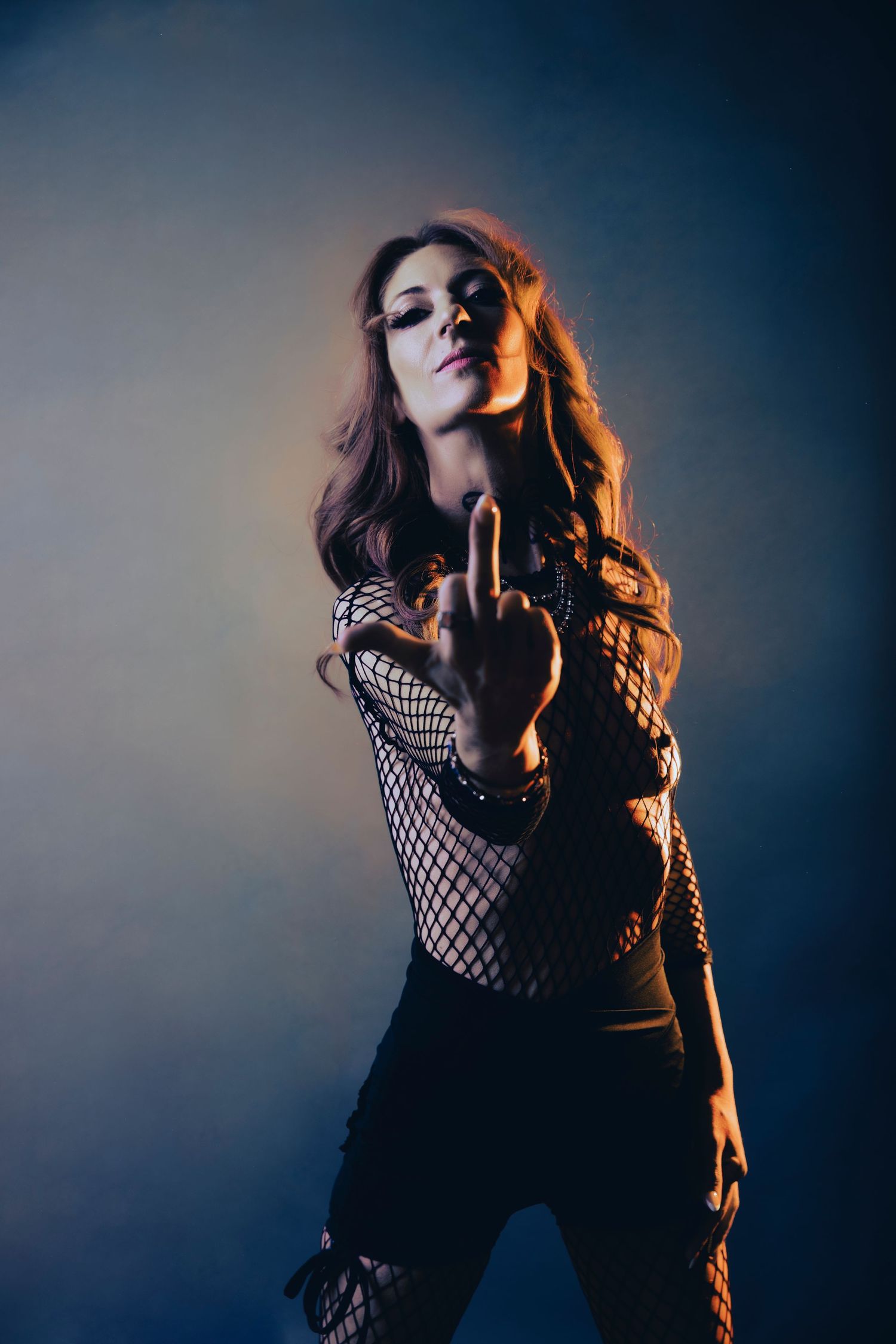 Woman in fishnet top making hand gesture under dramatic orange and blue studio lighting.