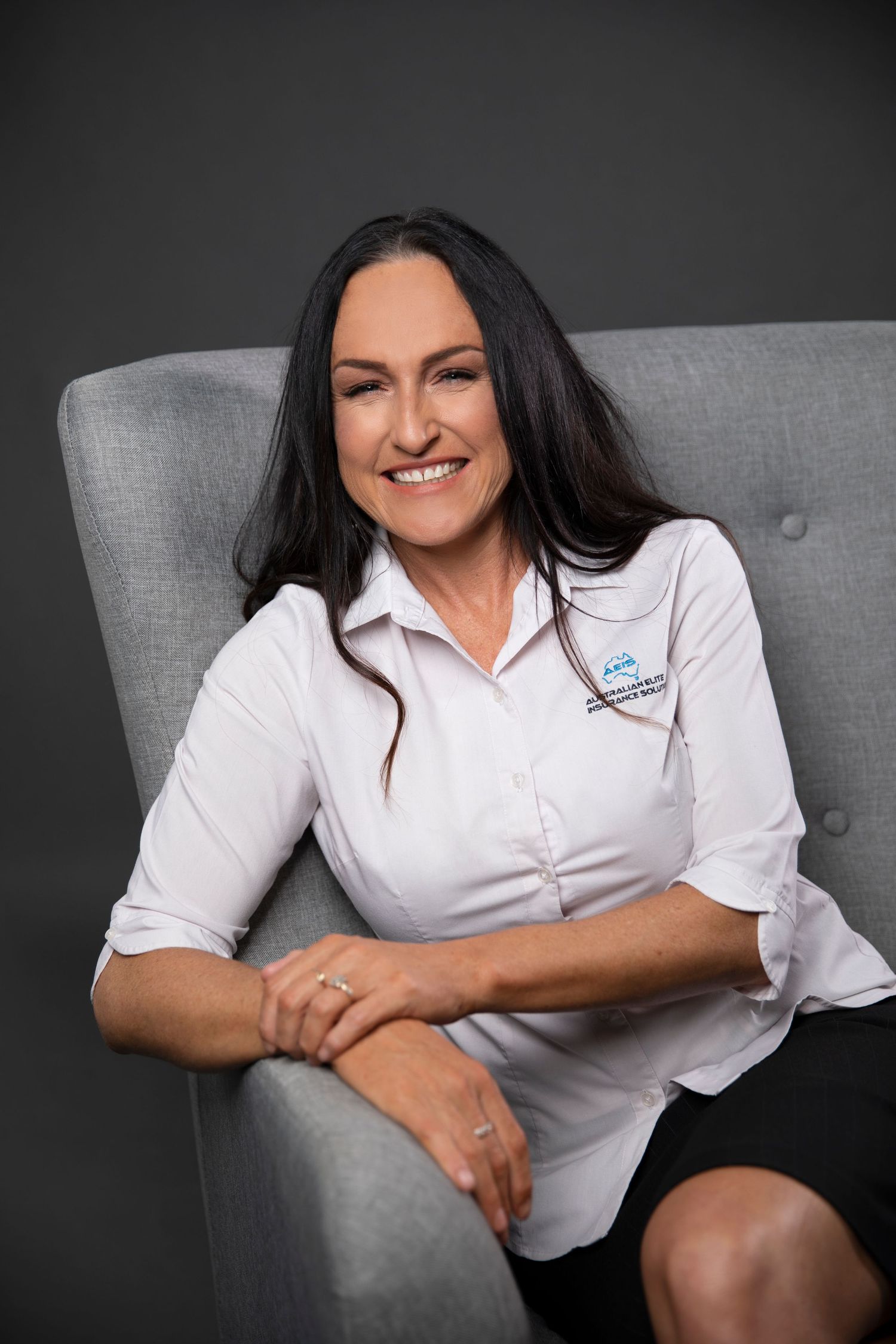 Confident woman with long dark hair in white business shirt sitting in gray armchair against dark background.