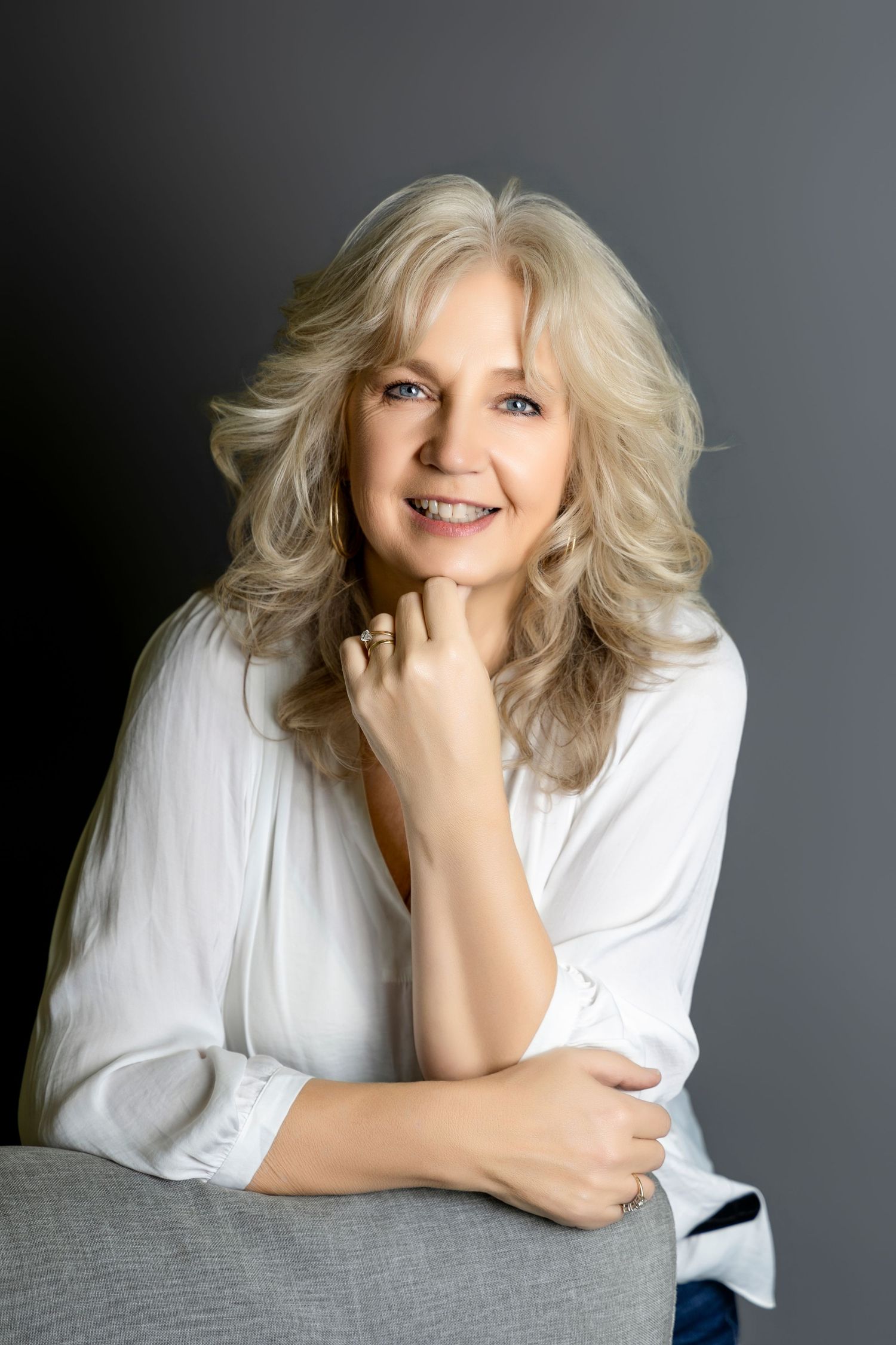 Smiling middle-aged blonde woman in white blouse poses relaxed against gray studio background.