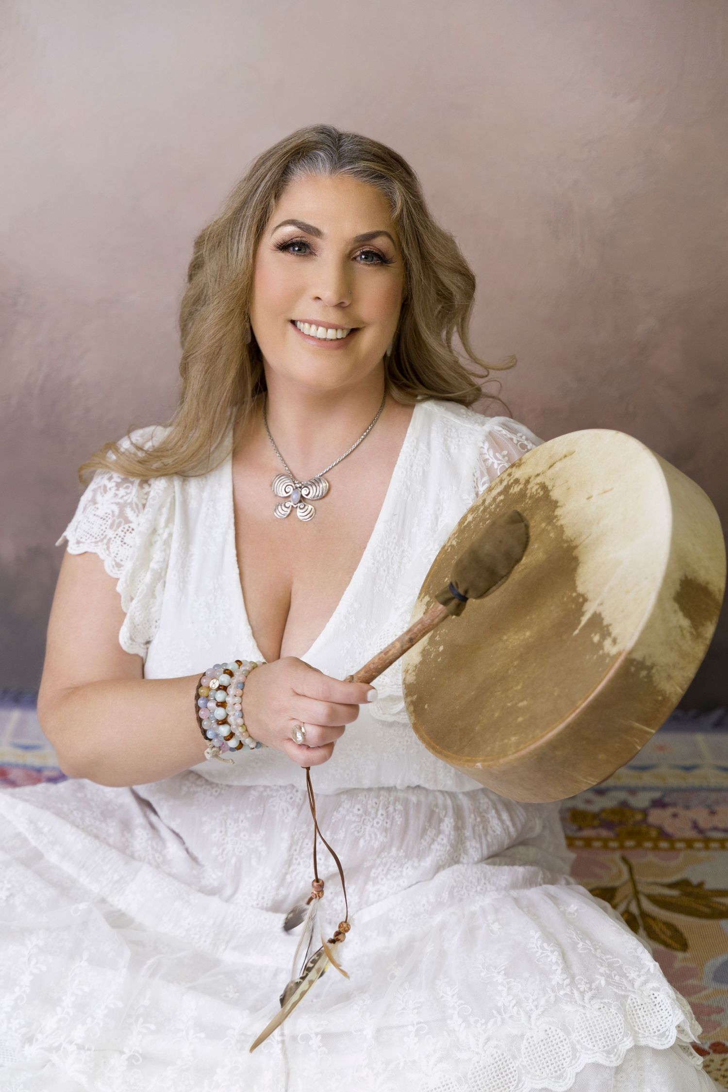 Smiling woman in white dress holding a shamanic drum in a professional portrait studio.