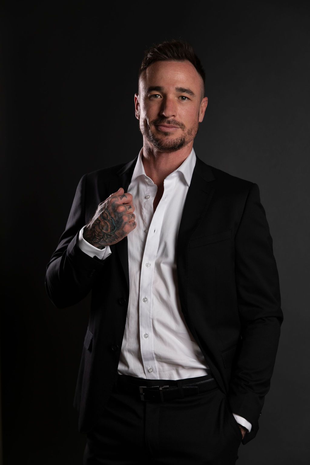 Tattooed man in black suit and white shirt posing confidently against dark background.