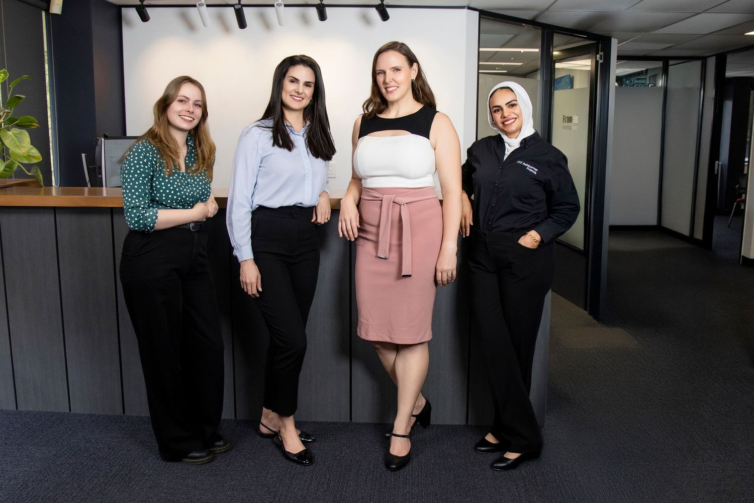 Four professional women standing together smiling in modern office corridor for team photo.
