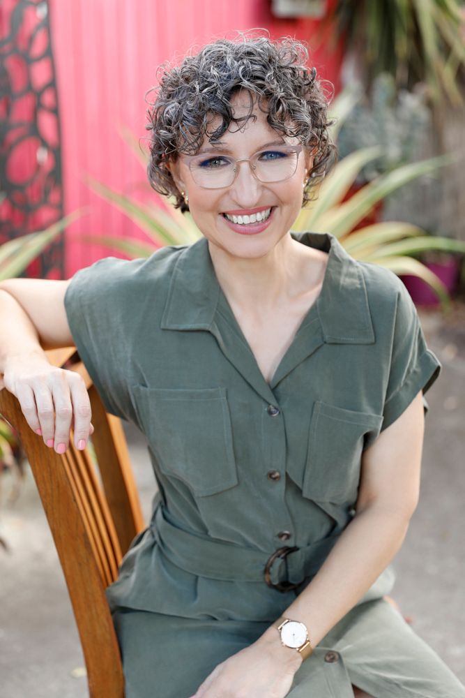 A curly-haired woman in green dress smiles while seated in a chair in an outdoor garden setting.