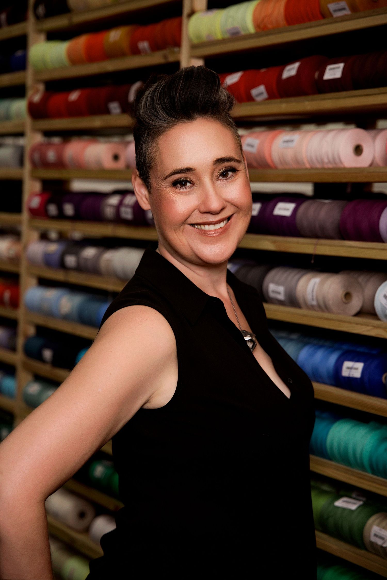 Smiling woman in black sleeveless top posing in front of shelves of colorful yarn spools in a craft store.