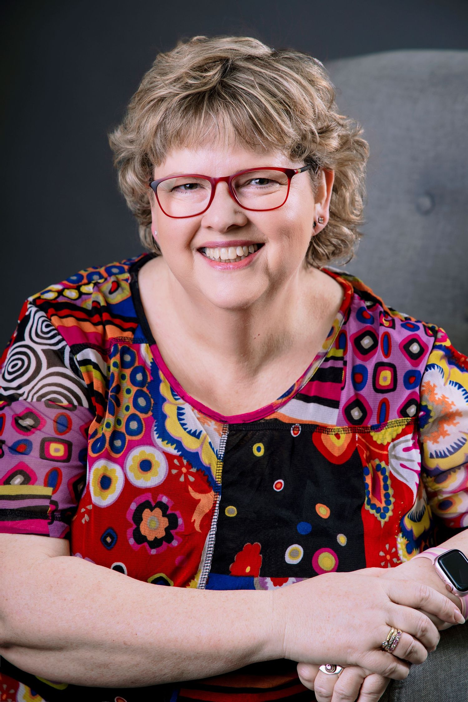 Cheerful woman with curly blonde hair and red glasses wearing a colorful patterned top seated in grey chair.