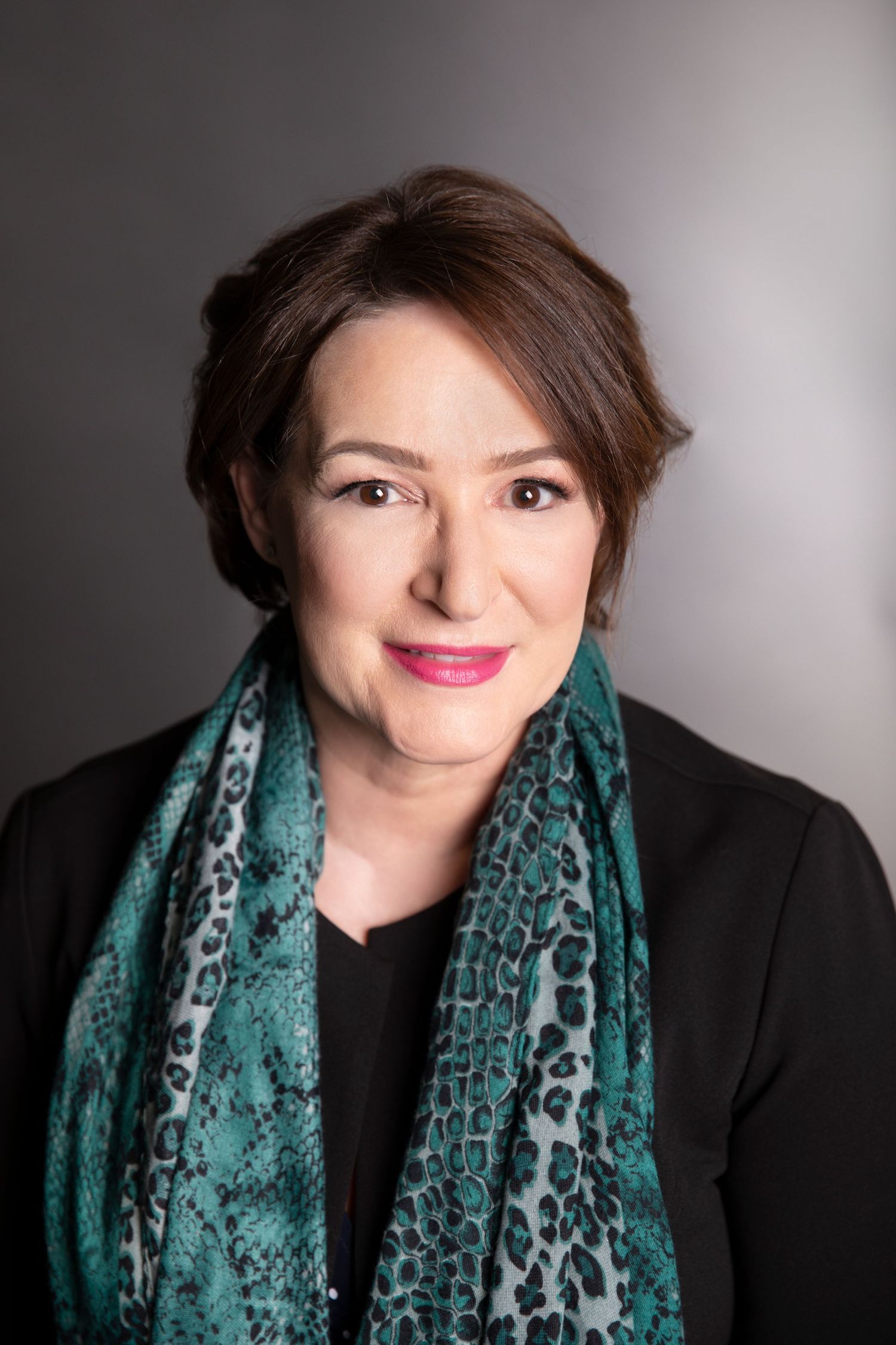 Professional woman with dark hair wearing a teal leopard print scarf and black blazer against grey background.