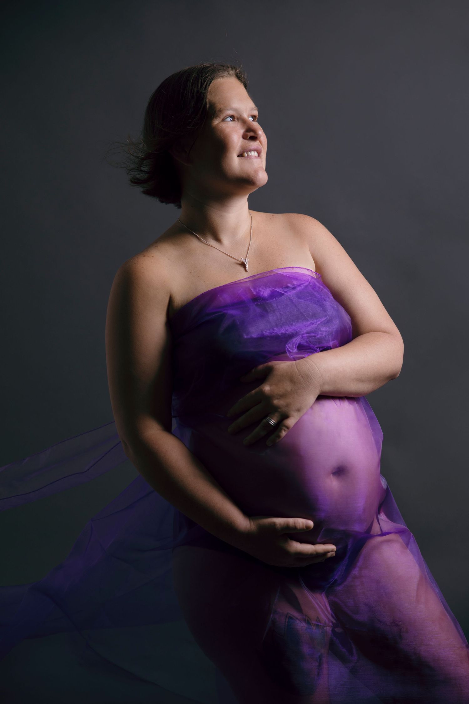 Pregnant woman wrapped in flowing purple fabric cradling baby bump against dark background.