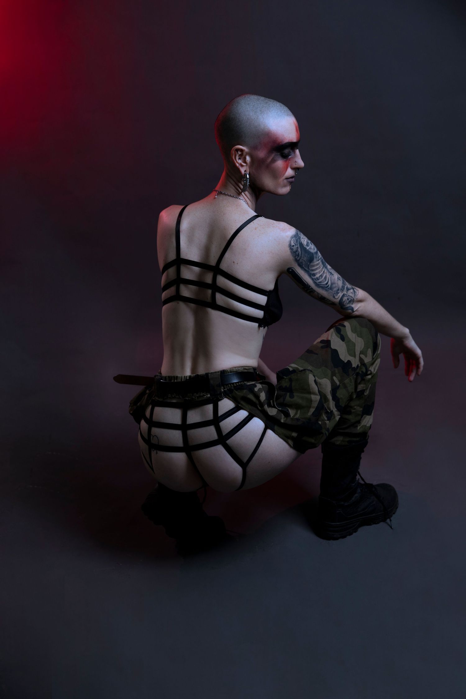 A tattooed bald woman in a strappy black harness top and camouflage pants poses in a dark studio.
