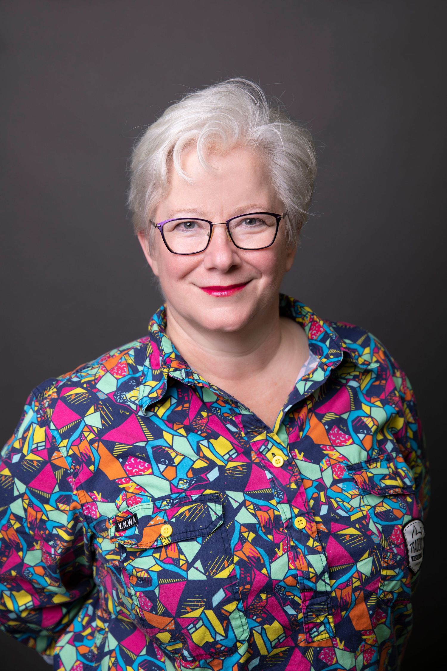 Cheerful senior woman with white hair and glasses in colorful patterned shirt portrait.