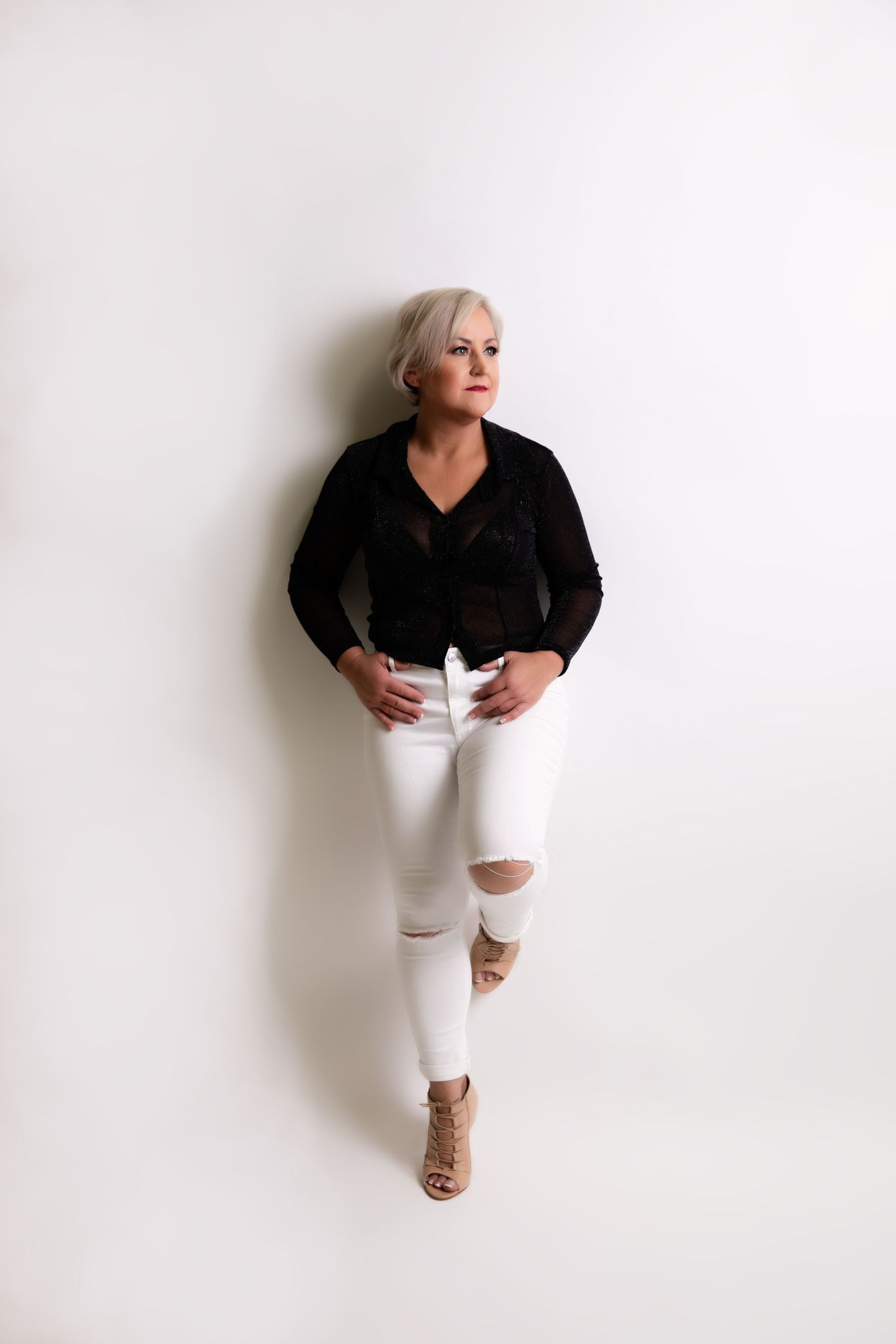 Stylish blonde woman in black top and white ripped jeans posing against white wall.
