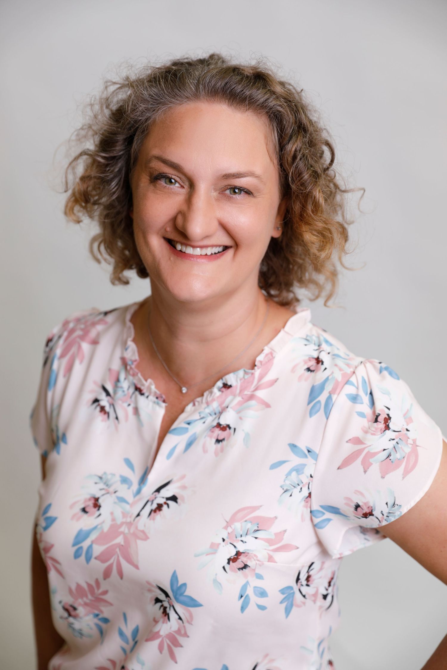 Happy woman with curly hair in floral blouse smiling against light gray background.