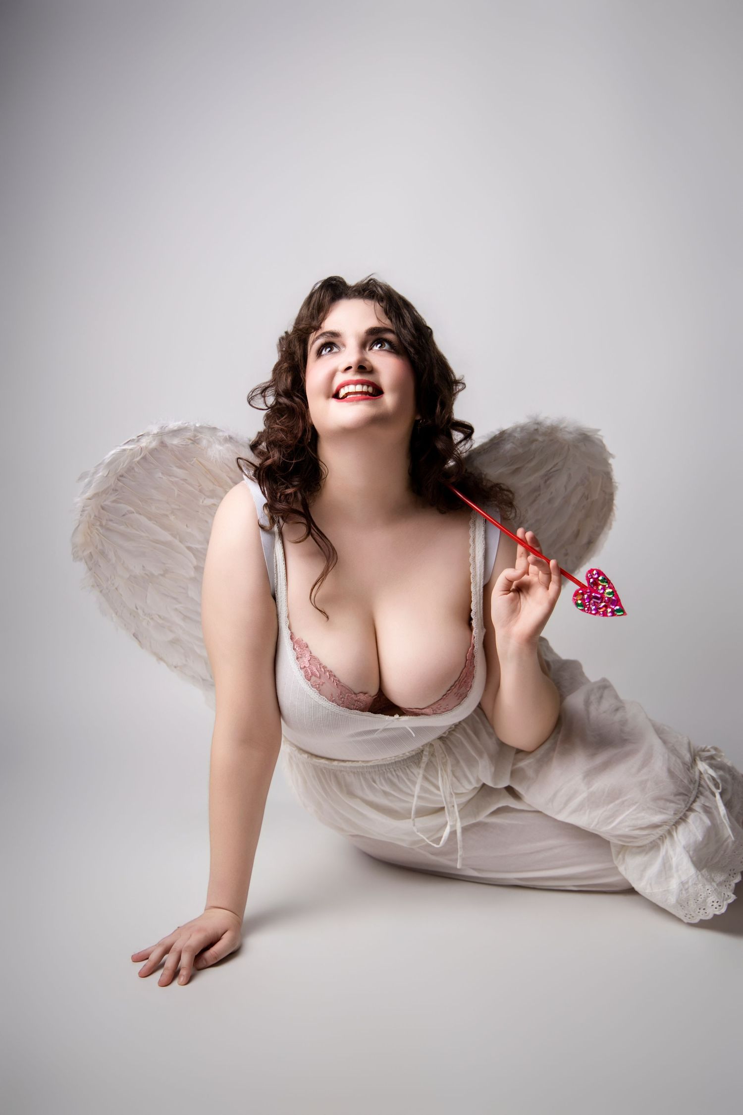 Brunette woman in angel costume with wings and pink bra holding heart arrow in studio photo.