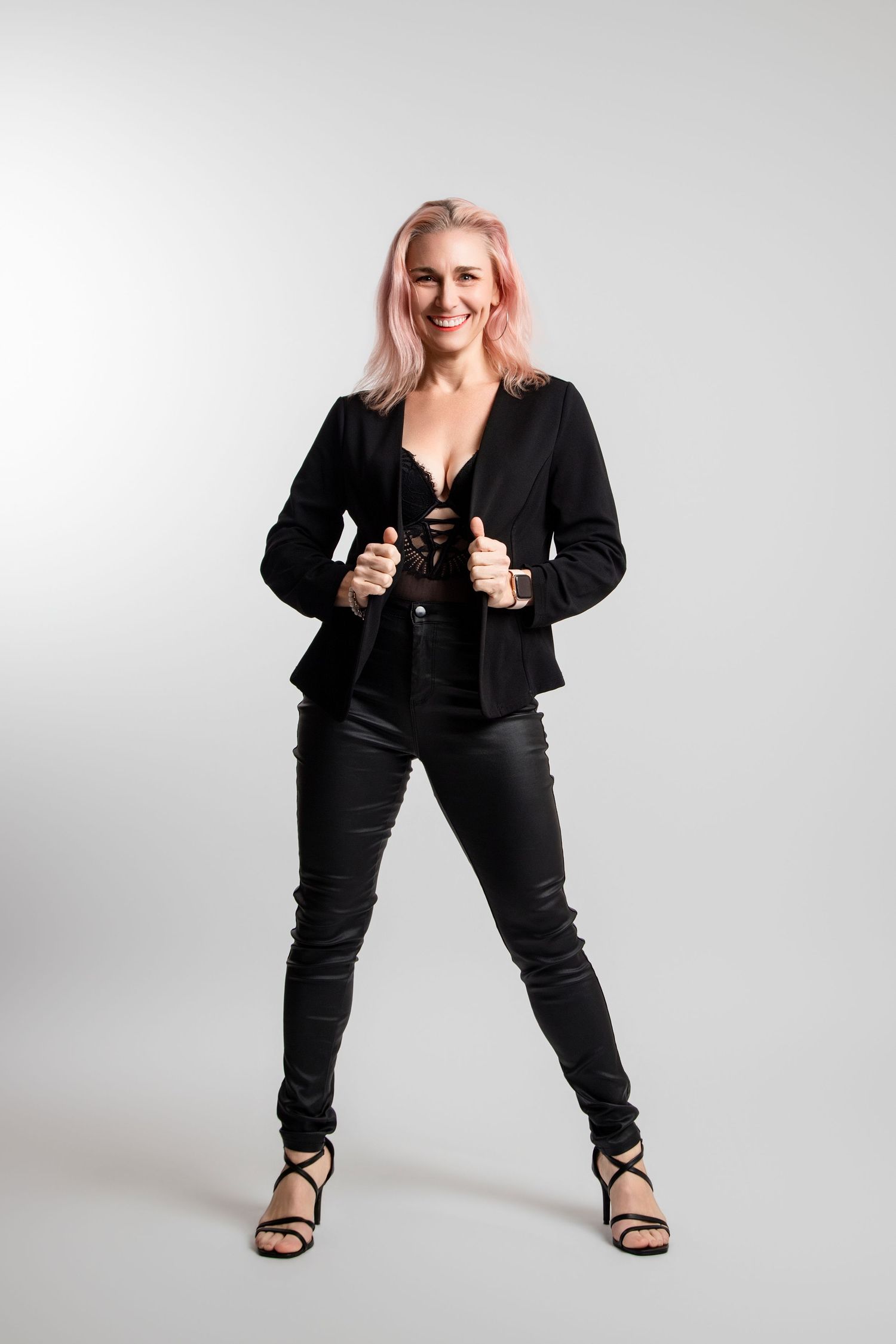 Smiling woman with pink hair in black blazer and leather pants posing in studio.
