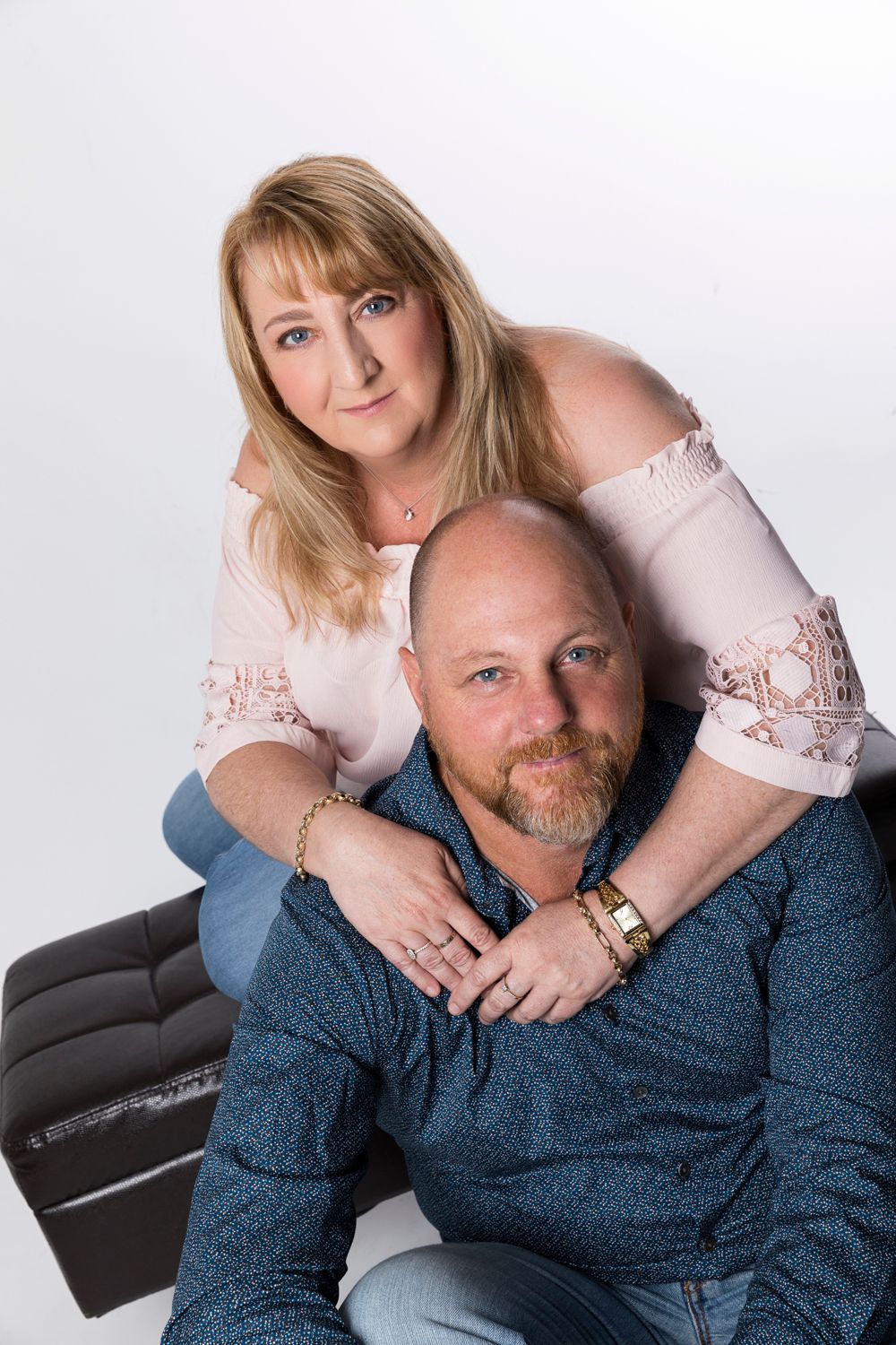 A blonde woman leans over a seated bearded man in a bright studio couples portrait photo.