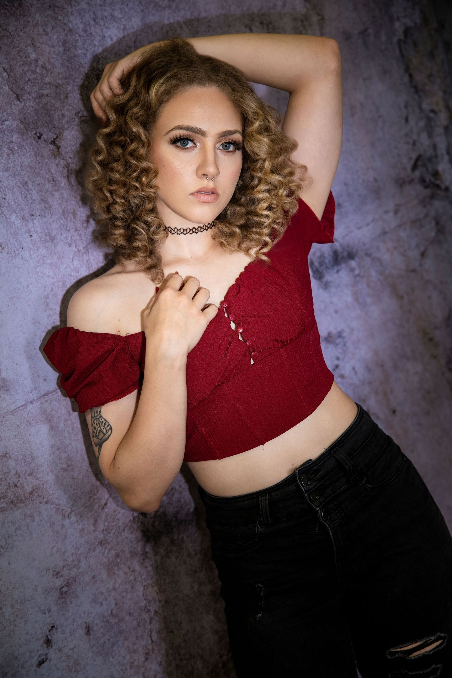 Young woman with curly blonde hair in red off-shoulder crop top against textured wall.