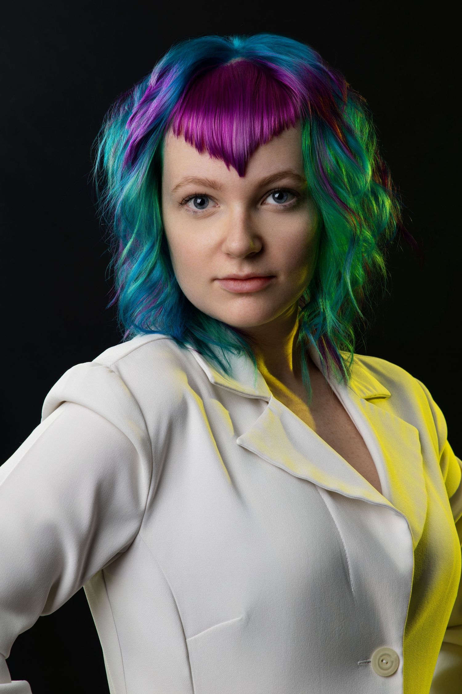 Woman with vibrant multicolored hair in white blazer posing against black background.
