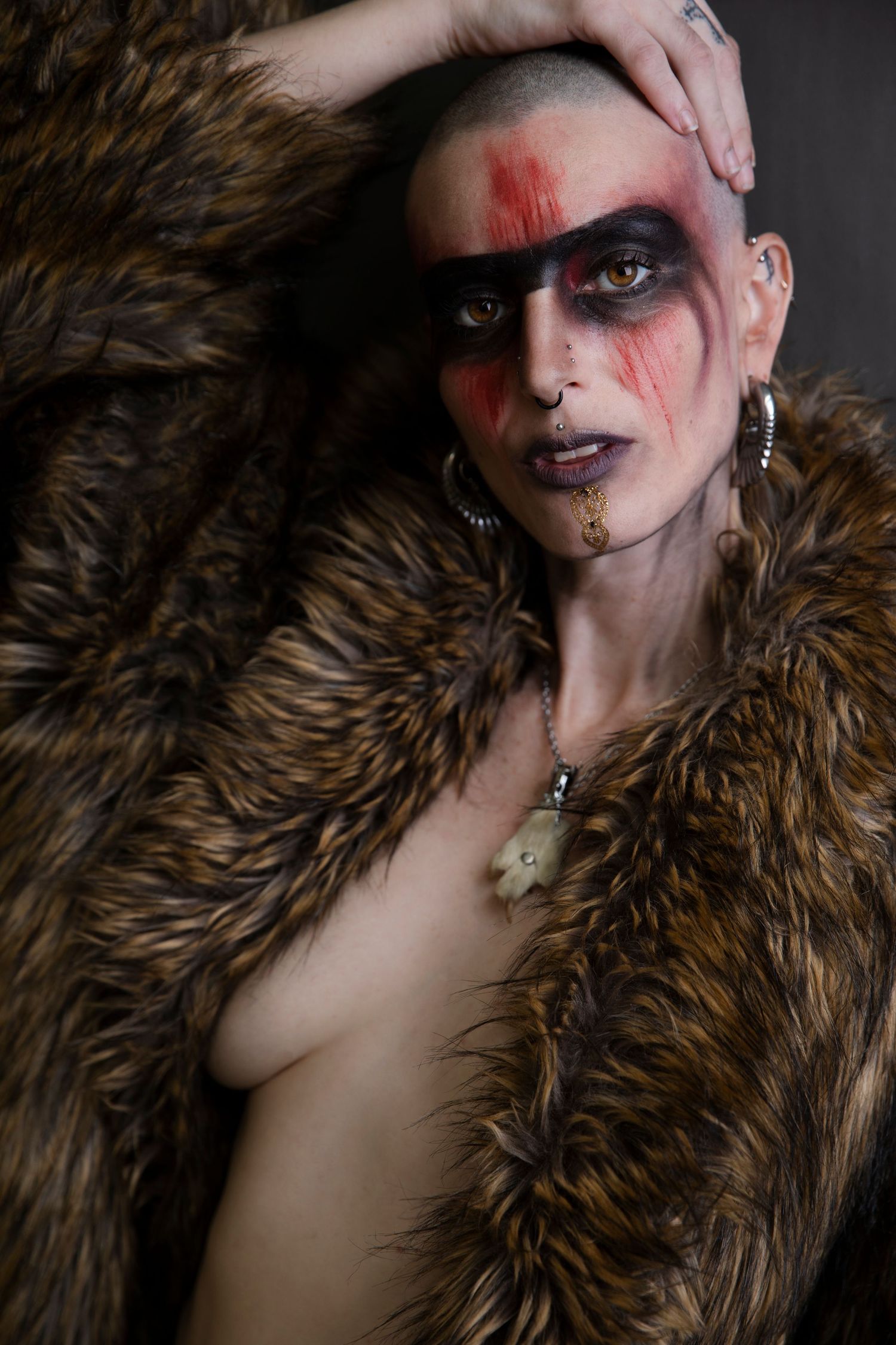 A bald woman with dramatic red and black face paint poses in an open fur coat in a studio.