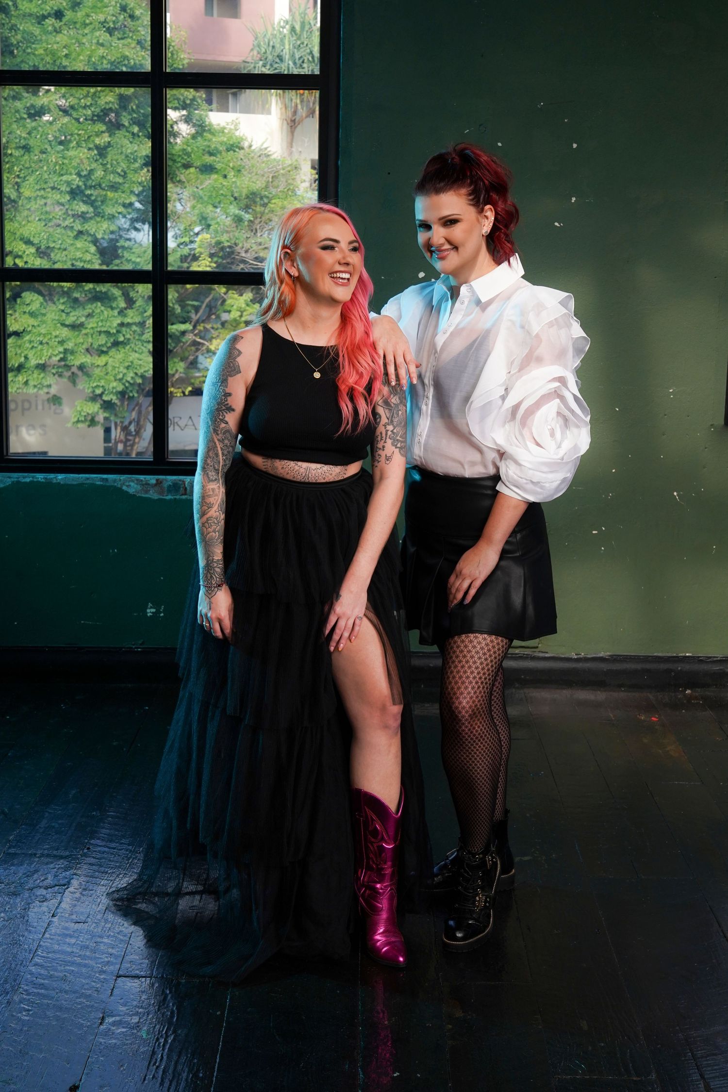 Two smiling women pose together in black and white outfits near large studio windows.