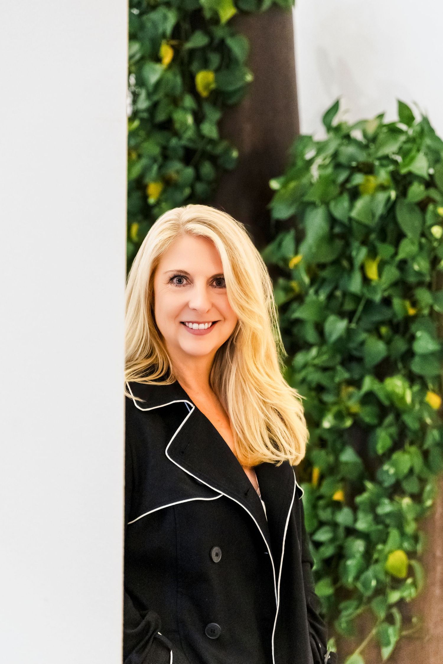 Smiling blonde woman in black coat peeking around a white pillar with green foliage behind.