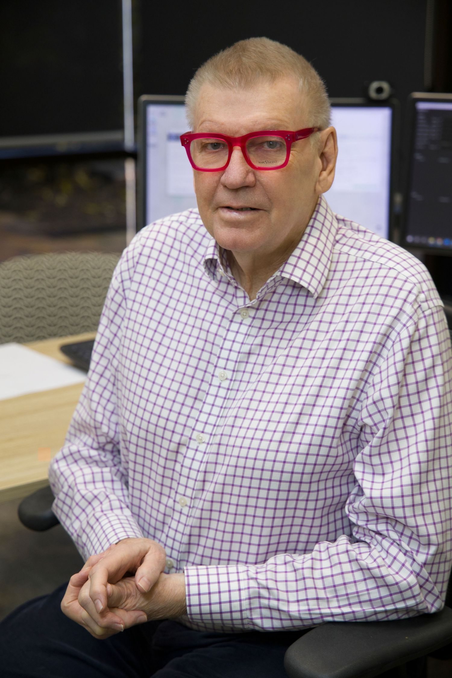 Senior man with red glasses in checkered shirt seated at desk with computer monitors in office setting.