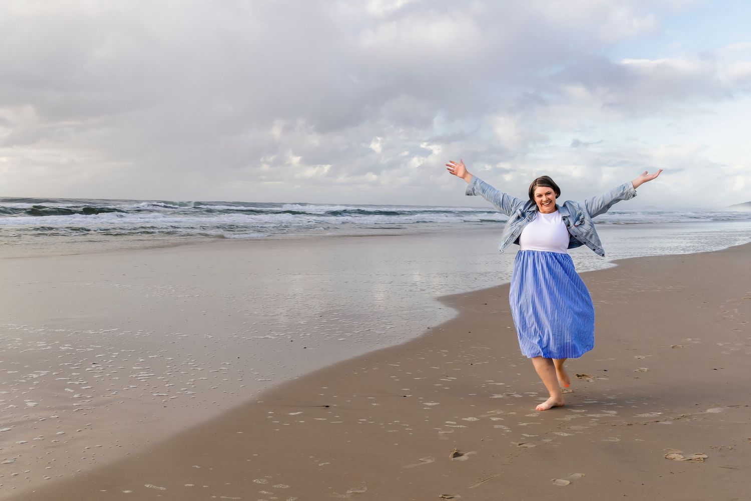 Woman in blue skirt and denim jacket joyfully posing on a sandy beach with arms outstretched.
