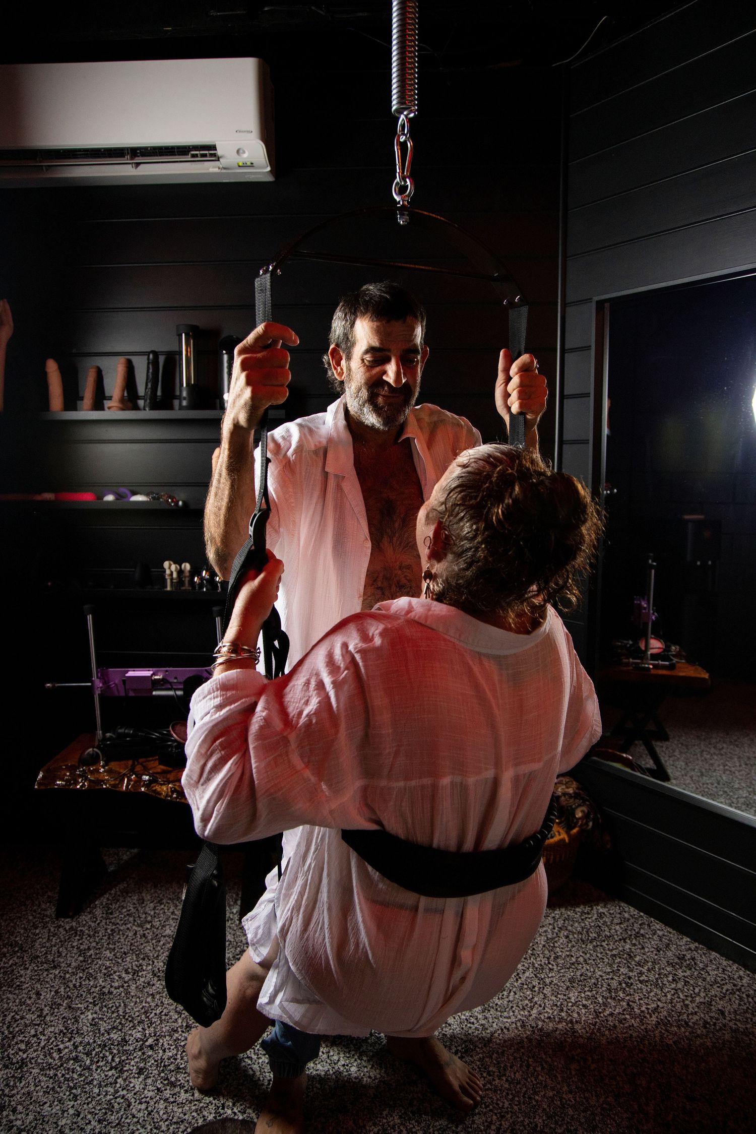 Two men in white robes in a dimly lit room with adult toys on shelves in the background.