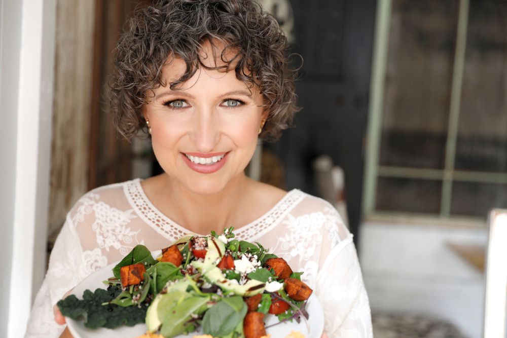 A smiling curly-haired woman holds a fresh colorful salad bowl outdoors.