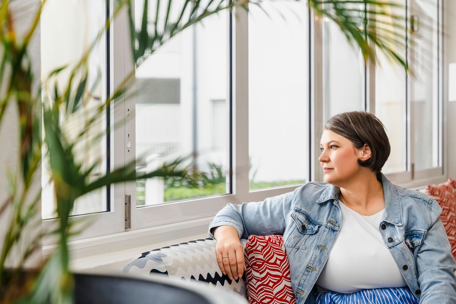 Woman in denim jacket sitting on a couch gazing out large windows near indoor plants.
