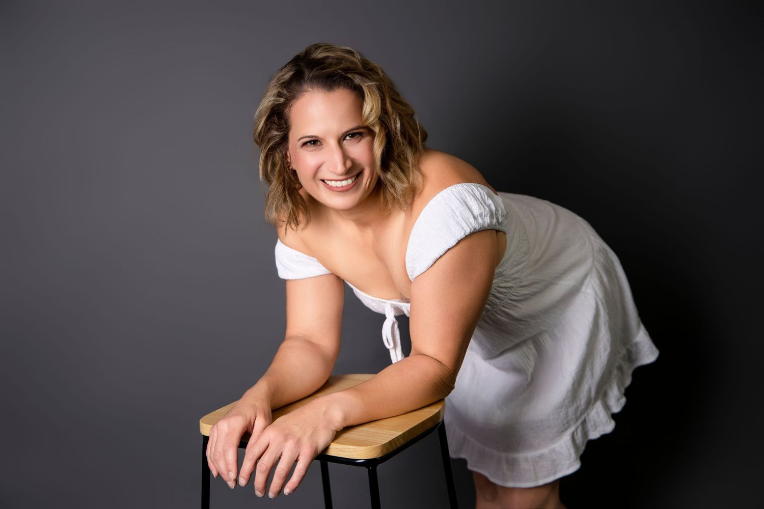 Smiling woman in white off-shoulder dress leaning on stool in studio.