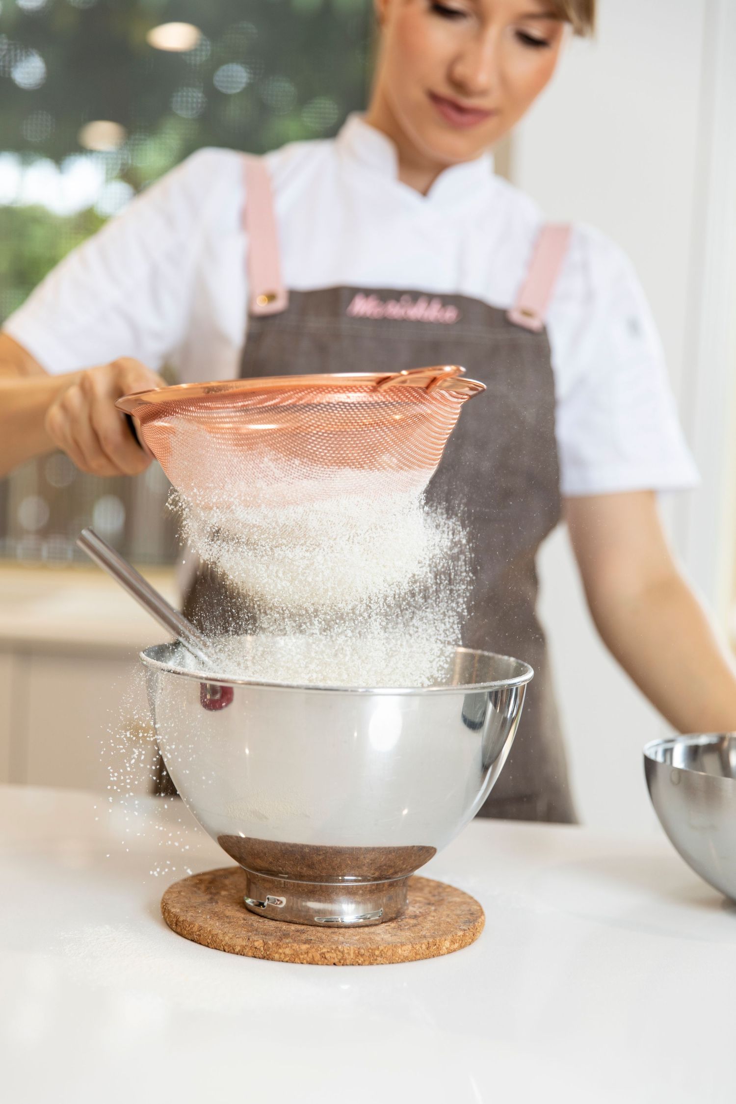 Female pastry chef in grey apron sifting flour into stainless steel mixing bowl in kitchen.